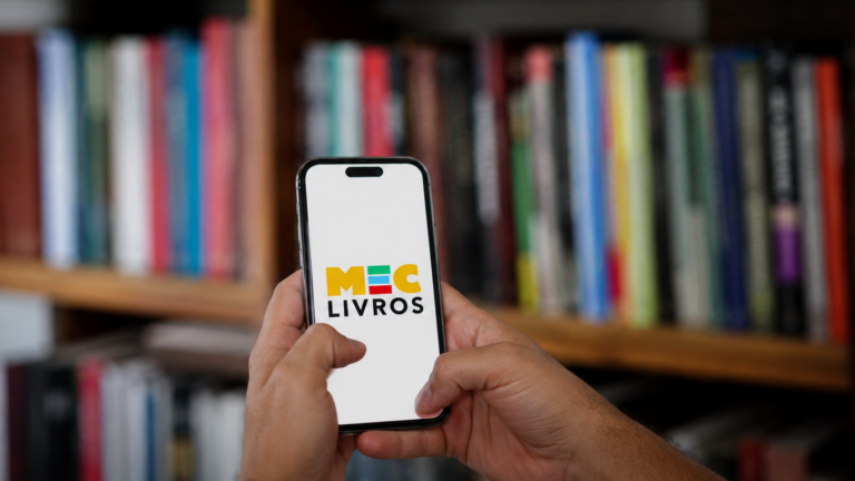 Pessoa segurando celular com a plataforma MEC Livros aberta diante de uma estante desfocada