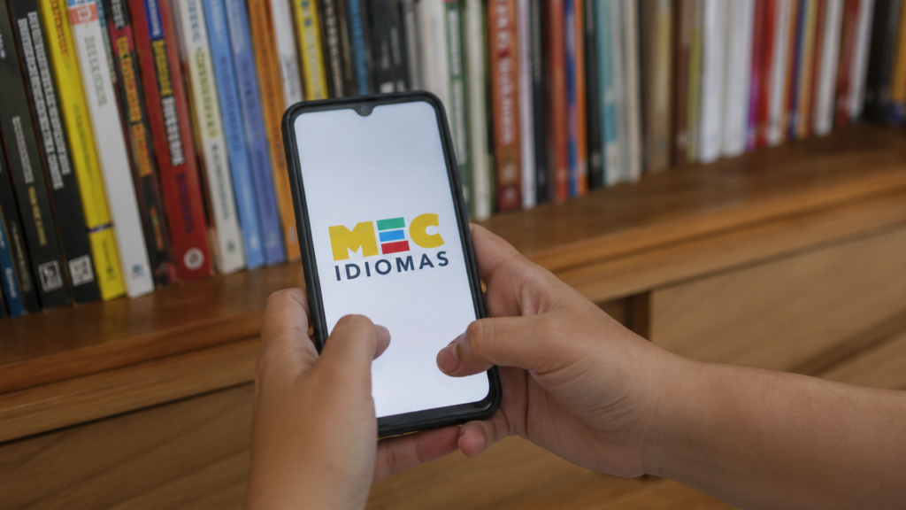 Pessoa segurando celular com a tela do MEC Idiomas diante de uma estante de livros